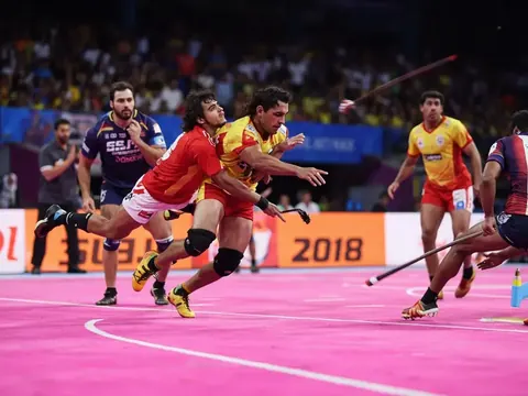 Pro Kabaddi raider mid-raid with defender lunging at ankle on a vivid pink mat